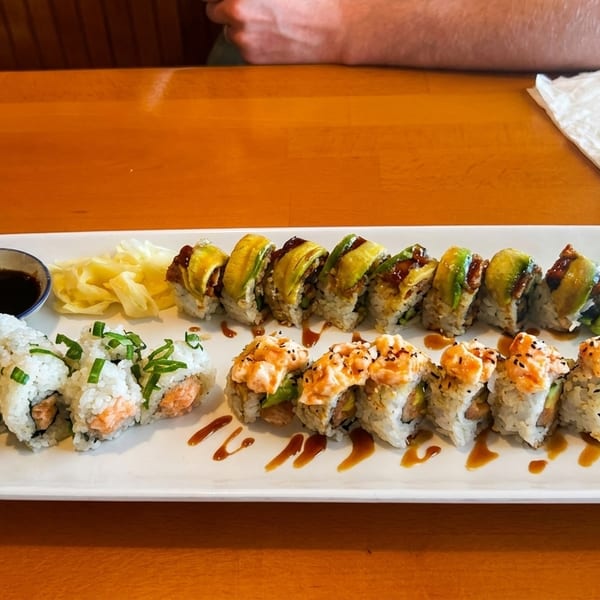 A white platter of three different sushi rolls, ginger, and soy sauce on a wooden table.