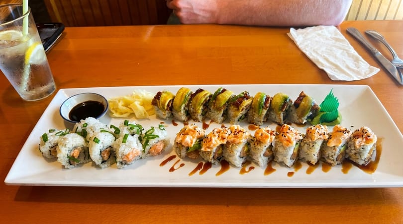 A white platter of three different sushi rolls, ginger, and soy sauce on a wooden table.