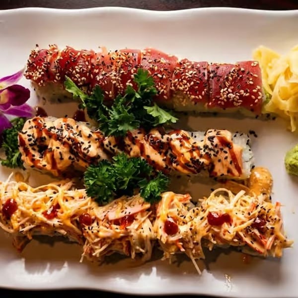 Three rows of sushi rolls with various toppings, ginger, wasabi, and an orchid on a white plate.