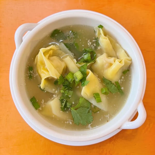 Wonton soup in a white bowl, garnished with green onions and cilantro on an orange table.