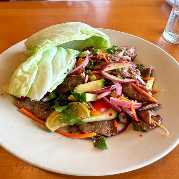 Spicy beef salad with red onions, cucumbers, carrots, and lettuce wraps on a white plate.