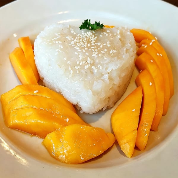 Heart-shaped sticky rice with sesame seeds and parsley, surrounded by sliced mango on a white plate.