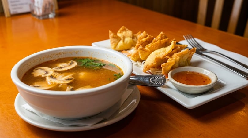 A bowl of mushroom soup and a plate of fried wontons with dipping sauce on a wooden table.