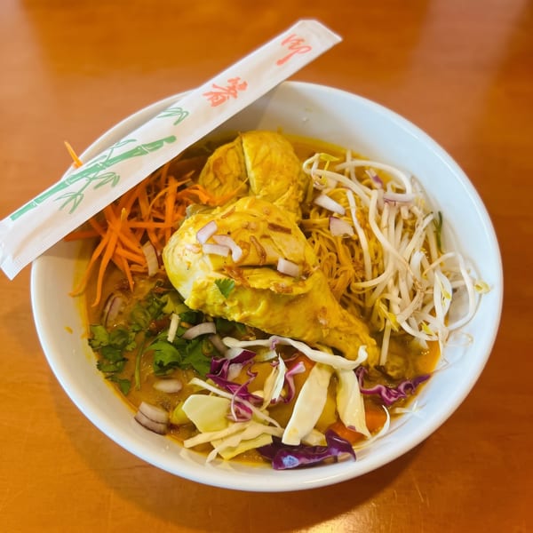 A bowl of yellow curry noodles with chicken, bean sprouts, carrots, and cabbage, with chopsticks.