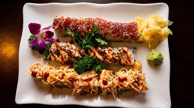 Three rows of sushi rolls with various toppings, garnished with a purple orchid, ginger, and wasabi.